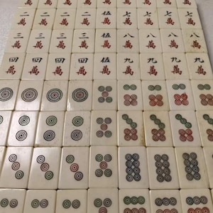 Puede incluir: Una colecci&oacute;n de fichas de mahjong rectangulares de color blanco roto dispuestas en una cuadr&iacute;cula. Las fichas presentan caracteres chinos grabados en tinta roja y negra, as&iacute; como patrones circulares en colores rojo, verde y negro.