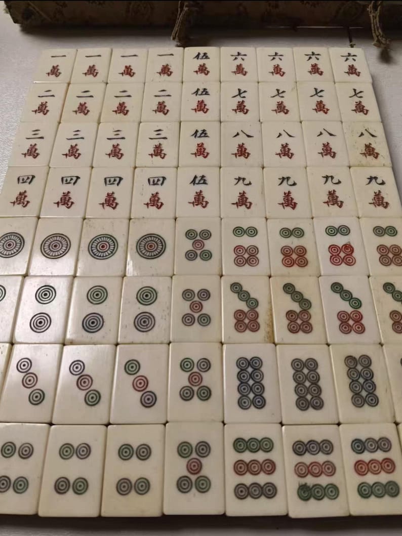Puede incluir: Una colecci&oacute;n de fichas de mahjong rectangulares de color blanco roto dispuestas en una cuadr&iacute;cula. Las fichas presentan caracteres chinos grabados en tinta roja y negra, as&iacute; como patrones circulares en colores rojo, verde y negro.