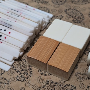 Puede incluir: Un conjunto de componentes de juego de Mahjong que incluye una pila de varillas de puntuaci&oacute;n blancas, largas y delgadas, con marcas negras y rojas, cuatro fichas rectangulares con superficies blancas y de madera, y dos dados blancos con puntos rojos y azules.