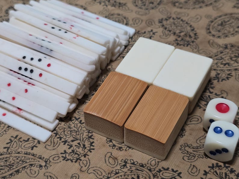 Puede incluir: Un conjunto de componentes de juego de Mahjong que incluye una pila de varillas de puntuaci&oacute;n blancas, largas y delgadas, con marcas negras y rojas, cuatro fichas rectangulares con superficies blancas y de madera, y dos dados blancos con puntos rojos y azules.