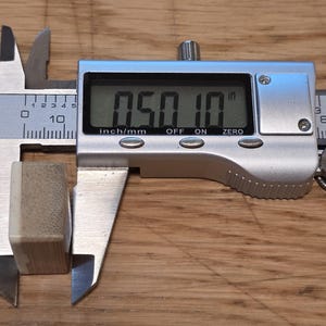 May include: A small, rectangular, light-colored block with a textured surface being measured by a silver digital caliper. The digital display on the caliper clearly shows a measurement of 0.50 inches, indicating the width of the object.