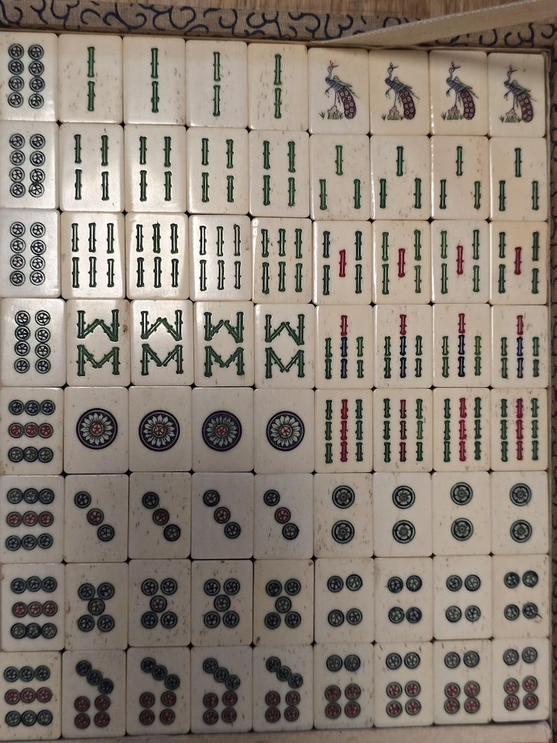 May include: A set of off-white Mahjong tiles arranged in a grid, featuring engraved symbols including green bamboo sticks, circular patterns, and bird illustrations. The tiles display various numerical and suit markings in green, red, and blue ink.