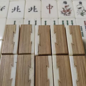 Puede incluir: Una colecci&oacute;n de fichas de Mahjong rectangulares dispuestas en filas. El primer plano presenta diez fichas con un dise&ntilde;o bicolor, que combina un material blanco liso con una veta de madera clara texturizada. El fondo muestra fichas blancas con caracteres de tinta negra y roja e ilustraciones florales.