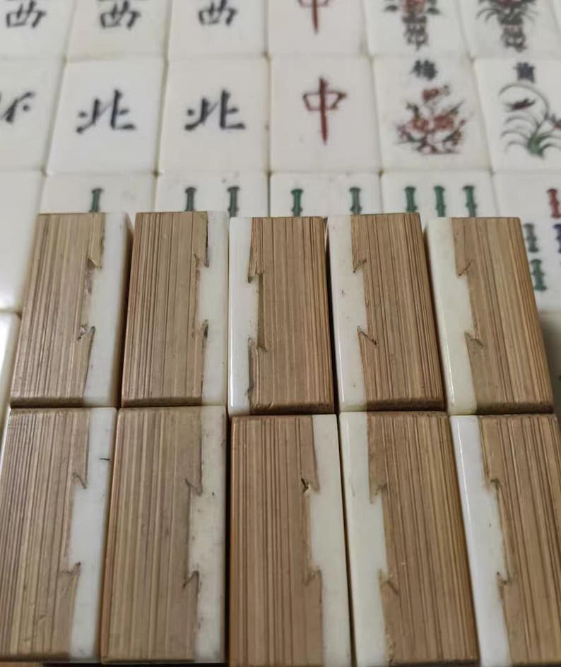 Puede incluir: Una colecci&oacute;n de fichas de Mahjong rectangulares dispuestas en filas. El primer plano presenta diez fichas con un dise&ntilde;o bicolor, que combina un material blanco liso con una veta de madera clara texturizada. El fondo muestra fichas blancas con caracteres de tinta negra y roja e ilustraciones florales.