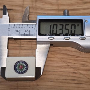 May include: A rectangular off-white mahjong tile featuring a circular floral design in green, blue, and red. The tile is positioned within a silver digital caliper that displays a measurement of 1.0350 inches on its clear liquid crystal screen.