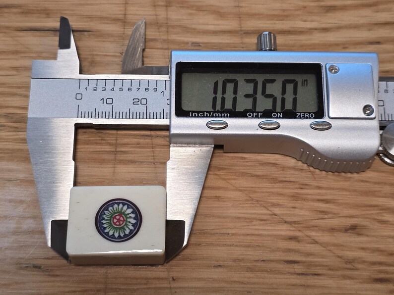 May include: A rectangular off-white mahjong tile featuring a circular floral design in green, blue, and red. The tile is positioned within a silver digital caliper that displays a measurement of 1.0350 inches on its clear liquid crystal screen.