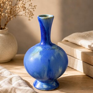 May include: A small, round ceramic vase with a narrow neck and a flared base. The vase features a vibrant blue glaze with lighter, cloud-like white and pale green streaks throughout the surface, creating a smooth, glossy, and variegated appearance.