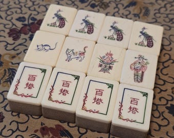May include: A collection of twelve off-white rectangular mahjong tiles arranged in three rows. The tiles feature engraved illustrations of peacocks, cats, a decorative urn, a figure in traditional robes, and the Chinese characters 百搭 in red ink.