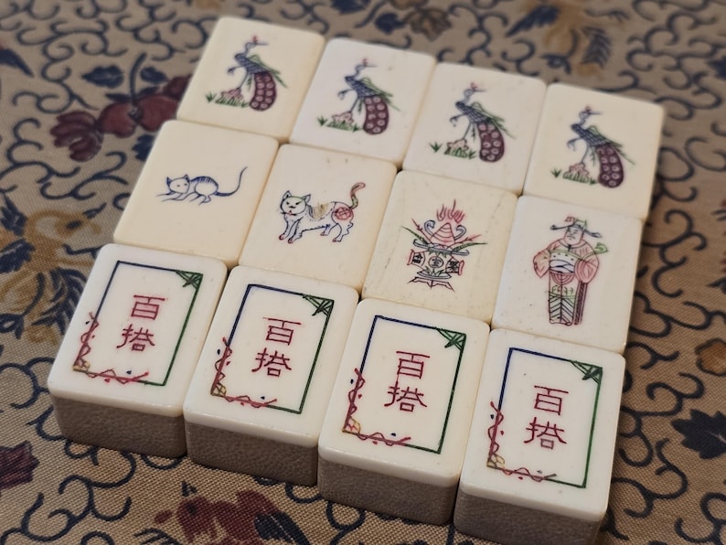 May include: A collection of twelve off-white rectangular mahjong tiles arranged in three rows. The tiles feature engraved illustrations of peacocks, cats, a decorative urn, a figure in traditional robes, and the Chinese characters 百搭 in red ink.