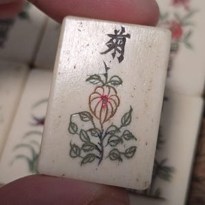 Puede incluir: Una ficha de Mahjong rectangular de color blanco hueso con una ilustraci&oacute;n grabada y pintada a mano de una flor con hojas verdes y un centro rojo. La parte superior muestra un car&aacute;cter chino negro y la superficie tiene textura.