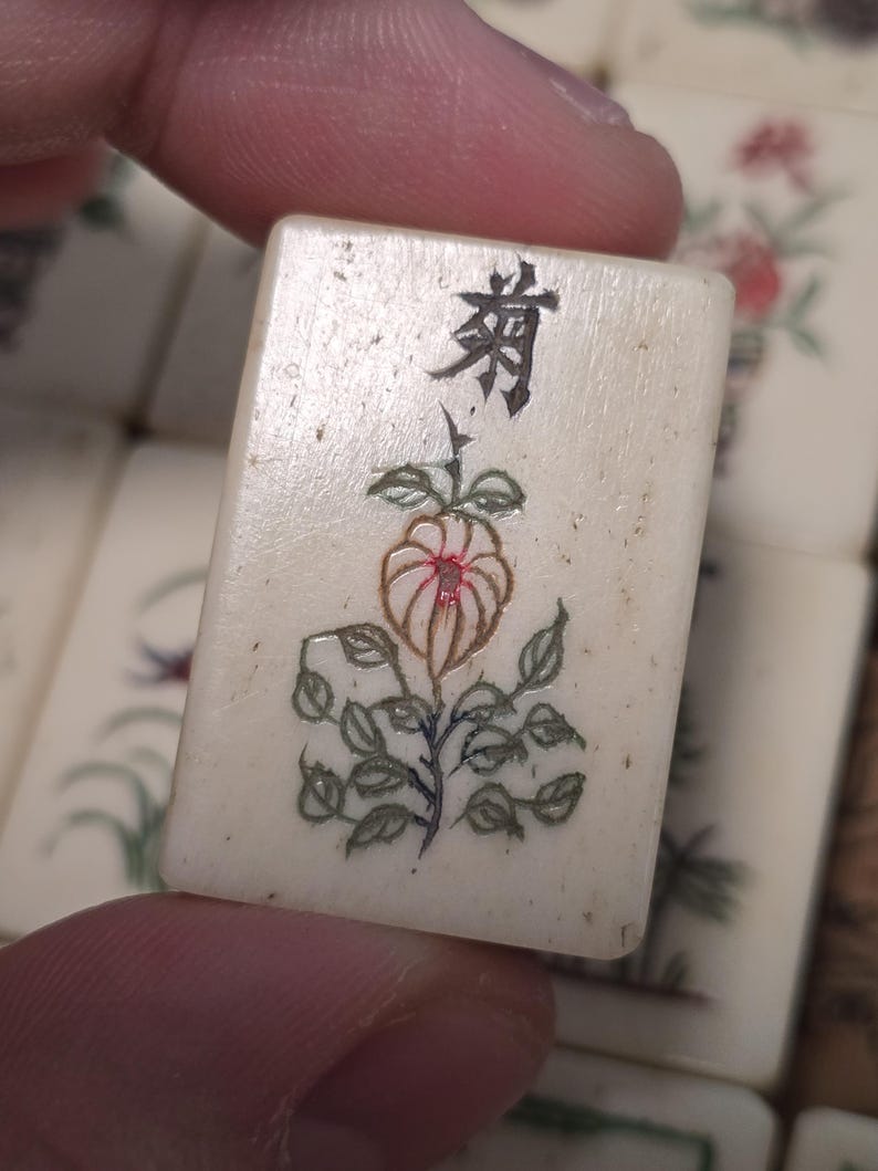 Puede incluir: Una ficha de Mahjong rectangular de color blanco hueso con una ilustraci&oacute;n grabada y pintada a mano de una flor con hojas verdes y un centro rojo. La parte superior muestra un car&aacute;cter chino negro y la superficie tiene textura.