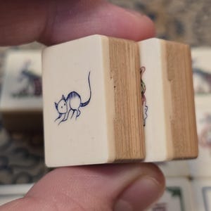 May include: Two rectangular mahjong tiles with a cream-colored front face and a light brown wooden back. The front of the left tile features a dark blue line drawing of a small mouse. The right tile shows a partial colorful graphic illustration.