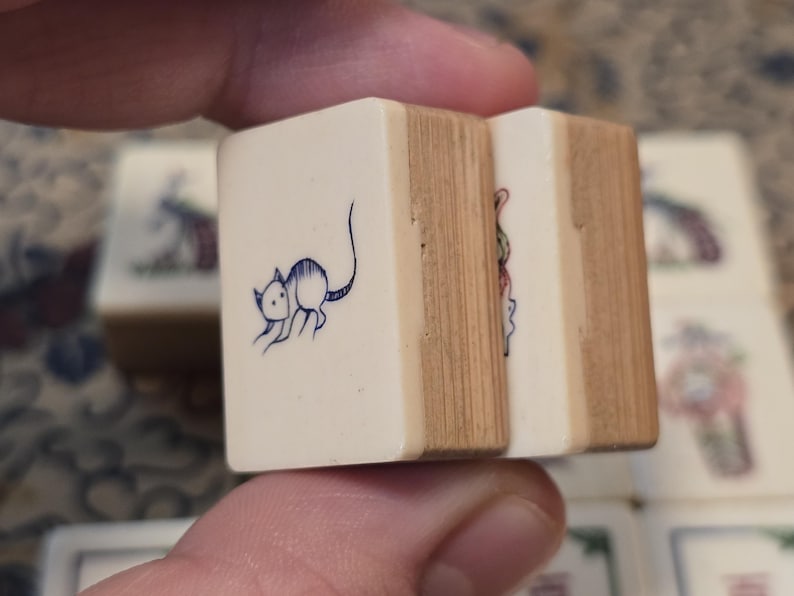 May include: Two rectangular mahjong tiles with a cream-colored front face and a light brown wooden back. The front of the left tile features a dark blue line drawing of a small mouse. The right tile shows a partial colorful graphic illustration.