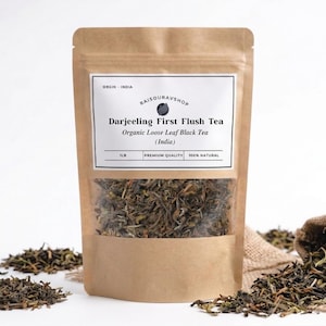 May include: A brown paper bag with a clear window displaying loose leaf tea. The label reads "Darjeeling First Flush Tea" and "Organic Loose Leaf Black Tea (India)". A small burlap sack and loose tea leaves are scattered around the bag.