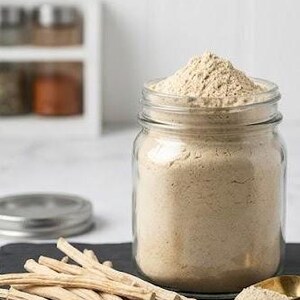 May include: A clear glass jar filled with light brown powder, topped with a mound of the same powder. A small gold spoon and a pile of light-colored sticks are next to the jar on a black surface. A few sticks are in the foreground.