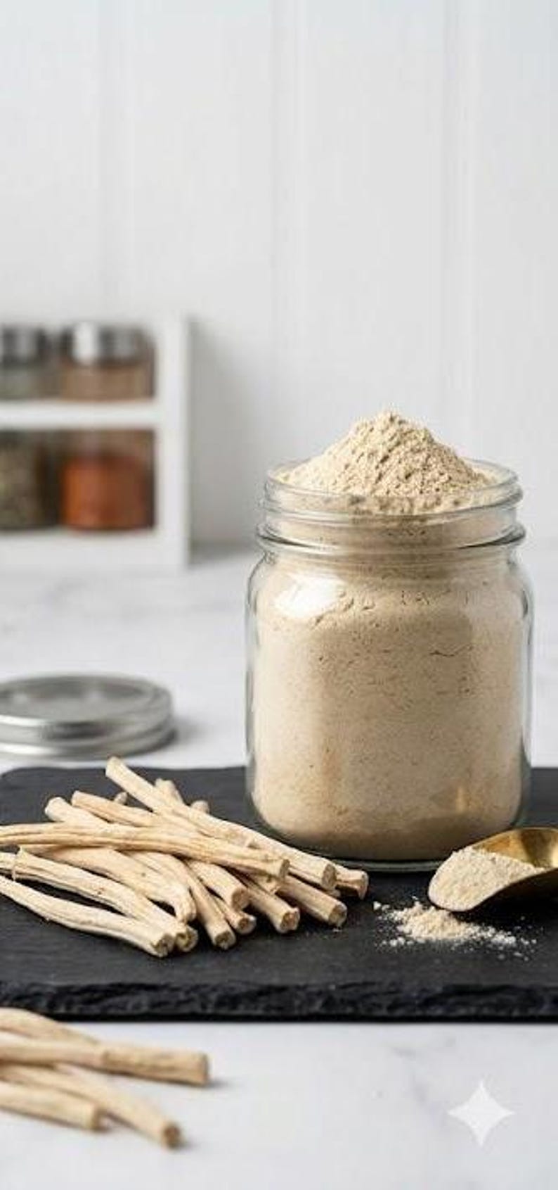 May include: A clear glass jar filled with light brown powder, topped with a mound of the same powder. A small gold spoon and a pile of light-colored sticks are next to the jar on a black surface. A few sticks are in the foreground.