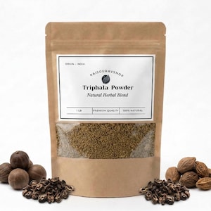 May include: A brown paper bag of Triphala Powder, a natural herbal blend, with a clear window showing the contents. The bag is labelled with the text "RAISOUBAYSHOP" and "1 LB". Several dried fruits and seeds are arranged around the bag.