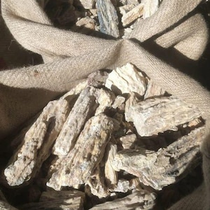 May include: A burlap sack filled with pieces of petrified wood. The wood pieces are various sizes and shapes, with a light brown and beige color. The texture of the wood appears rough and weathered.