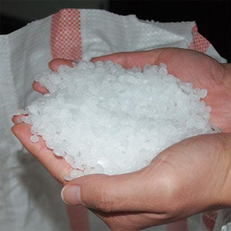 May include: A large quantity of small, translucent white wax pellets held in a cupped shape. The pellets are circular, flat, and uniform in size, appearing as a pile of raw material granules intended for melting or crafting purposes.