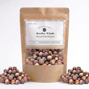 May include: A brown paper bag with a clear window displaying Reetha Whole Natural Dried Soapnut. The bag has a white label with text and is surrounded by small piles of the soapnuts. The bag is labeled as 1 LB.