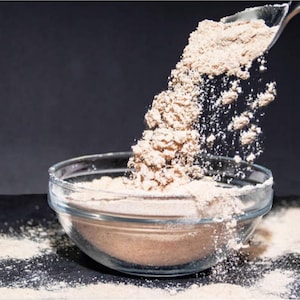 May include: A clear glass bowl filled with a light brown powder, with more powder being poured in from a metal spoon. The powder is spilling out of the bowl onto a dark surface, creating a dynamic image.