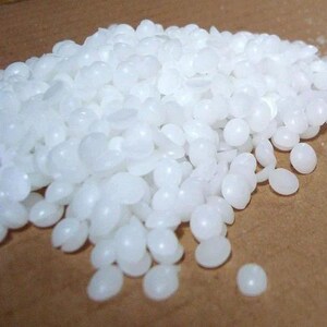 May include: A pile of small, round, white wax pellets. Each individual pellet has a smooth, opaque surface and a slightly flattened, circular shape. The collection of pellets is clustered together, creating a textured, granular appearance.