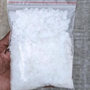 May include: A clear plastic resealable bag filled with small, irregular, translucent white crystalline granules. The bag features a distinct red plastic seal at the top, providing a secure closure for the contents contained within the transparent pouch.