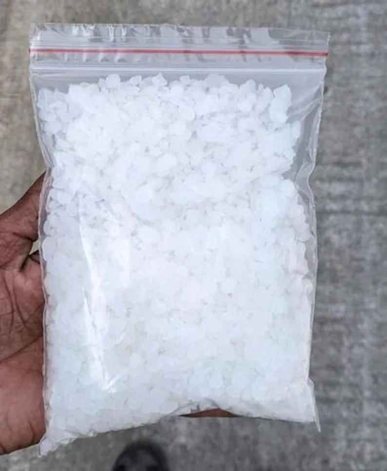 May include: A clear plastic resealable bag filled with small, irregular, translucent white crystalline granules. The bag features a distinct red plastic seal at the top, providing a secure closure for the contents contained within the transparent pouch.