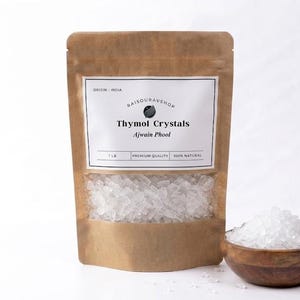 May include: A brown paper bag with a white label displaying "Thymol Crystals" and "Ajwain Phool". The bag contains clear crystals and is next to a small wooden bowl filled with similar crystals. The text "1 lb" is visible.
