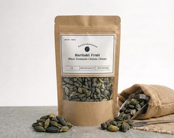 May include: A brown paper pouch containing dried Haritaki fruit, labeled with the text Haritaki Fruit Whole Terminalia Chebula (Dried) 1LB. A small pile of the dark, textured fruit sits in front, with more spilling from a burlap sack.