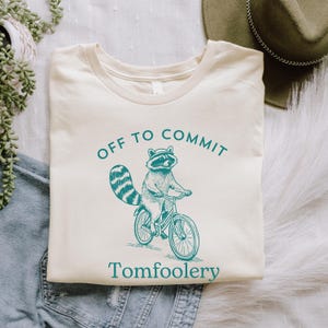 May include: A beige t-shirt featuring a graphic of a raccoon riding a bicycle. The text "Off to Commit Tomfoolery" is printed on the shirt.