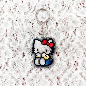 Può includere: Un portachiavi pixelato con un gatto bianco con un fiocco rosso, una tuta blu e un papillon giallo. Il portachiavi ha un anello e una catena argentati. Lo sfondo è un tessuto di pizzo bianco.