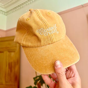 May include: A yellow baseball cap with the text "Custard Cream" embroidered in white on the front.