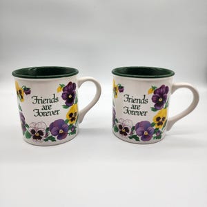 Op de afbeelding: Twee witte keramische mokken met een groene rand en oor. De mokken zijn versierd met paarse, gele en witte viooltjes en de tekst "Friends are Forever".