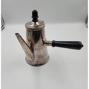 Puede incluir: Una cafetera plateada con un asa negra y un pomo negro en la tapa. La cafetera tiene un pico largo y un borde decorativo alrededor de la base y la tapa. La cafetera es probablemente de metal.