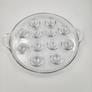 May include: Clear glass escargot plate with 12 individual wells. The plate is round with two small handles on either side. The wells are evenly spaced around the plate. The plate is made of clear glass.