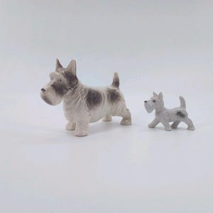 Peut inclure: Deux figurines en céramique de Scottish Terrier. Le plus grand chien est blanc avec des marques marron et se tient debout. Le plus petit chien est gris avec une tache sombre et marche. Les deux ont des détails.