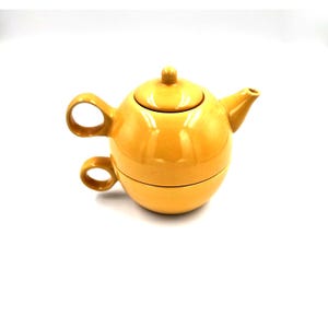 May include: A yellow ceramic teapot and teacup set. The teapot has a rounded shape with a lid and spout. The teacup is integrated into the base of the teapot, with a handle. The set is a solid, warm yellow color.
