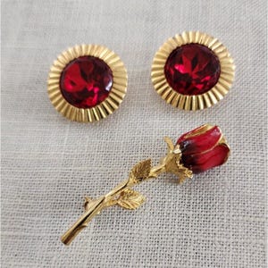 May include: A set of jewelry featuring a pair of gold-toned stud earrings with large, faceted red gems and a gold-toned rose brooch. The rose brooch has red enamel petals and gold leaves.