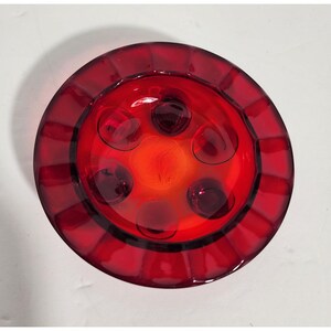 May include: A vibrant red glass ashtray with a circular design. The ashtray features a series of raised, rounded indentations and a central, lighter-colored area. The rim has a ribbed texture.