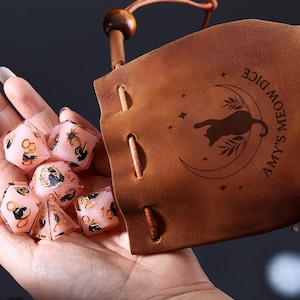 Pode incluir: Dados rosa com detalhes dourados e s&iacute;mbolos pretos, perto de um saco de couro castanho. O saco apresenta uma silhueta de gato numa lua crescente e o texto "Amy's Meow Dice".