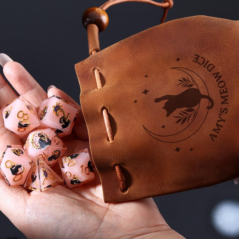 Pode incluir: Dados rosa com detalhes dourados e s&iacute;mbolos pretos, perto de um saco de couro castanho. O saco apresenta uma silhueta de gato numa lua crescente e o texto "Amy's Meow Dice".