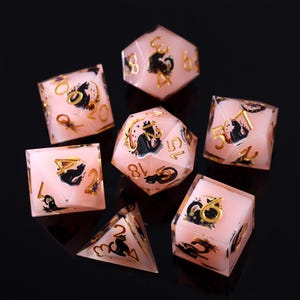 Pode incluir: Um conjunto de sete dados poli&eacute;dricos em v&aacute;rias formas. Os dados s&atilde;o rosa transl&uacute;cido com numera&ccedil;&atilde;o dourada e ilustra&ccedil;&otilde;es pretas de um gato. Os dados s&atilde;o para jogos de RPG.