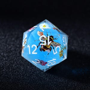 May include: A clear, multi-sided die with a bee and floral design suspended in a blue resin. The die features white numbers and is suitable for tabletop gaming. The bee is brown and black.