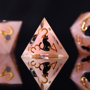 Puede incluir: Un dado transparente rosa de cuatro caras con números dorados y una silueta de gato negro. El dado se refleja en una superficie negra, con otros dados visibles en el fondo. Los números son 2, 3 y 4, con un símbolo de triángulo.