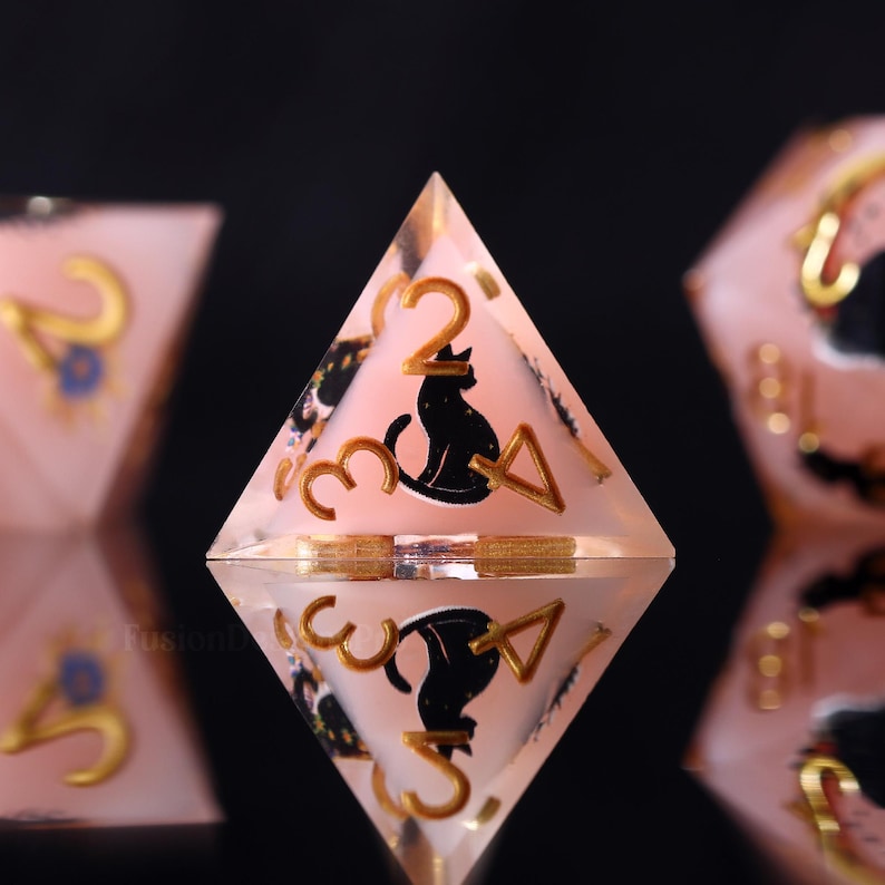 Pode incluir: Um dado transparente rosa de quatro lados com algarismos dourados e uma silhueta de gato preto. O dado &eacute; refletido em uma superf&iacute;cie preta, com outros dados vis&iacute;veis ao fundo. Os n&uacute;meros s&atilde;o 2, 3 e 4.