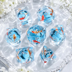 May include: Set of seven clear resin dice with a blue interior and orange koi fish inclusions. Each die features silver numbering. The dice are arranged on a clear, textured surface, with white flowers in the background.