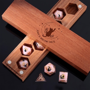 Könnte beinhalten: Holzbox mit der Aufschrift "Amy's Meow Dice" und einer Katzen-Silhouette. Sie enthält mehrere rosa Würfel mit schwarzen Katzenillustrationen und goldenen Zahlen. Die Würfel haben verschiedene Formen, darunter ein Würfel, eine Pyramide und ein 20-seitiger Würfel.