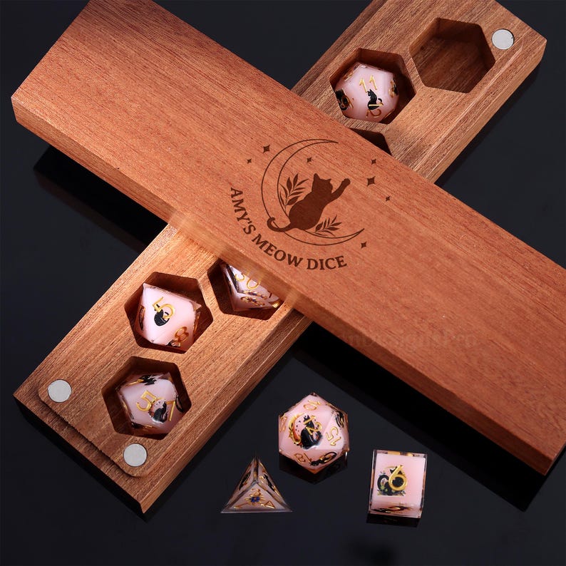 Pode incluir: Caixa de dados de madeira com o texto "Amy's Meow Dice" e uma silhueta de gato. A caixa cont&eacute;m v&aacute;rios dados rosa com ilustra&ccedil;&otilde;es de gatos pretos e numera&ccedil;&atilde;o dourada. Os dados t&ecirc;m v&aacute;rias formas, incluindo um cubo, uma pir&acirc;mide e um dado de 20 lados.