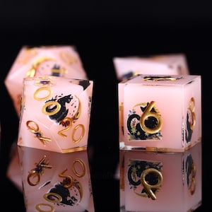 Pode incluir: Dados rosa e dourados com ilustra&ccedil;&otilde;es pretas e douradas. Os dados t&ecirc;m n&uacute;meros e s&iacute;mbolos dourados. Os dados est&atilde;o sobre uma superf&iacute;cie refletora.